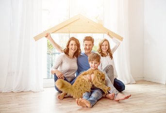 Glückliche Familie mit zwei Kindern und Katze sitzt im hellen Wohnzimmer und hält ein Holzhausdach über sich.