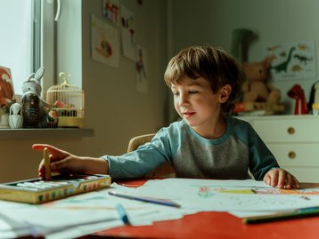 Junge zeichnet am Tisch in einem hellen Kinderzimmer mit Spielzeug und bunten Stiften.