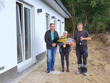 Feierliche Hausübergabe eines weißen, schlüsselfertigen Bungalows mit Blumenstrauß, Sekt und symbolischem Holzschlüssel an die Bauherrin