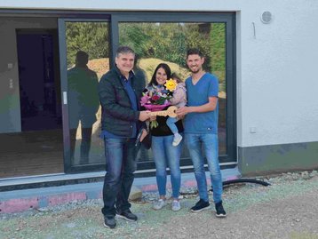 Persönliche Schlüsselübergabe des schlüsselfertigen Massivhauses: Bauleiter übergibt symbolischen Holzschlüssel an die junge Familie vor der Hebe-Schiebetür