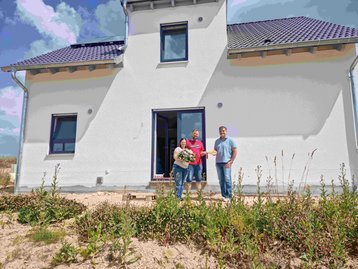 Schlüsselübergabe für das schlüsselfertige Massivhaus Flair 134: Bauherrenpaar mit Blumenstrauß und Bauleiter vor der Terrassentür des Neubaus